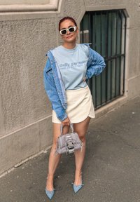 Giacca di jeans sopra una maglietta grafica blu chiaro, gonna avvolgente bianca, borsa grigia testurizzata e décolleté blu a punta.