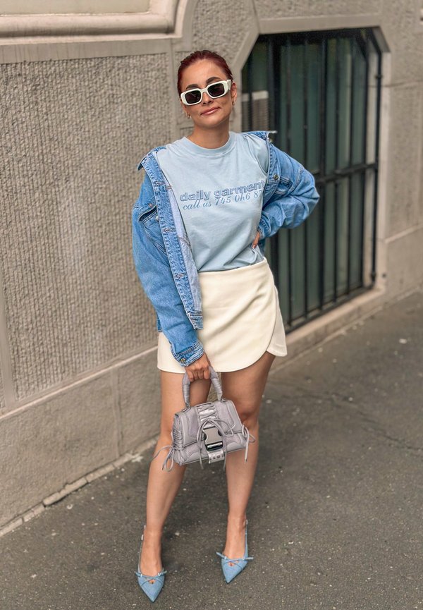 Giacca di jeans sopra una maglietta grafica blu chiaro, gonna avvolgente bianca, borsa grigia testurizzata e décolleté blu a punta.