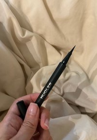 Hånd holder en sort flydende eyelinerpen med "MUGLER" label mod en beige stofbaggrund.