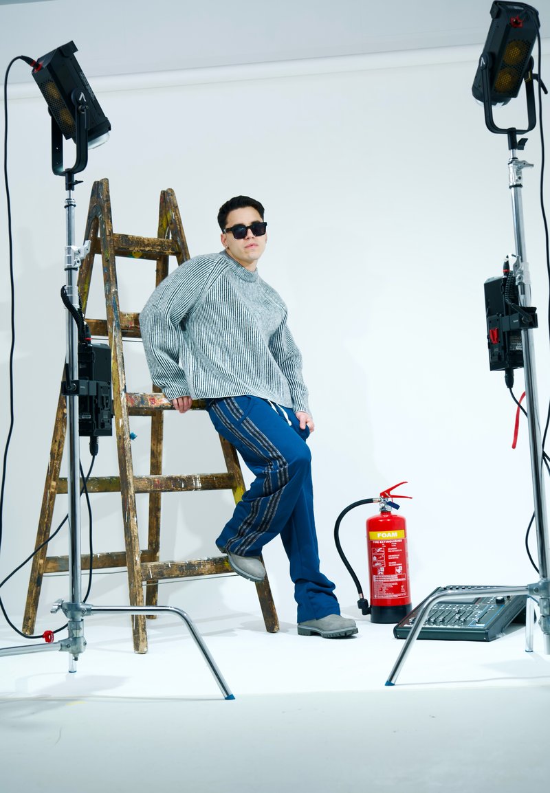 Grau gestreifter Strickpullover, blaue Hosen mit vertikalen Streifen, graue Schuhe, Sonnenbrille. Leiter und Feuerlöscher in einem Studio-Setting.