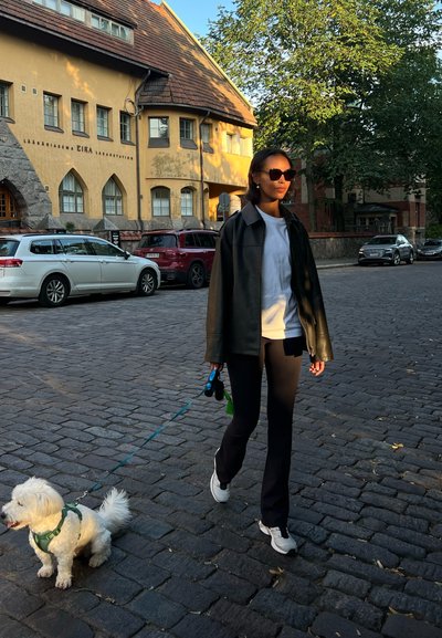 Una mujer pasea a un pequeño perro blanco con correa. Lleva una chaqueta de cuero negra, una camisa blanca, pantalones negros y zapatillas blancas.