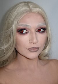 Porträt einer Person mit langen, welligen, platinblonden Haaren, roten Sklerakontaktlinsen und neutraler Lippenfarbe; die Merkmale umfassen dramatische Wimpern und Augenbrauen.