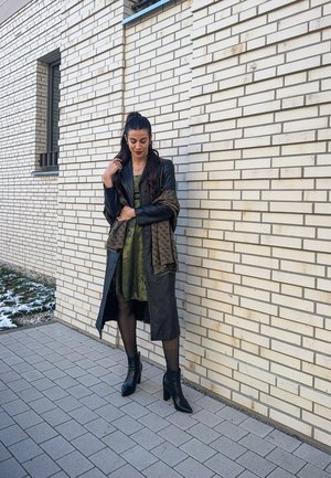 Largo abrigo de cuero negro sobre un vestido de satén verde, combinado con un chal ornamentado. Botines negros y medias con textura complementan el look.