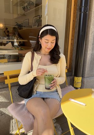 Una mujer con un cárdigan amarillo y una camiseta sin mangas está sentada sobre un cojín rosa en una silla de metal amarilla, sosteniendo una bebida verde con una pajita.