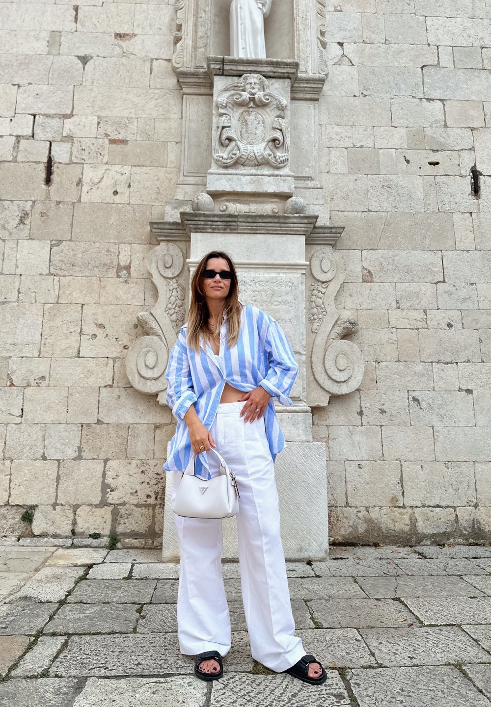 Camicia oversize a righe blu e bianche, pantaloni bianchi a gamba larga, sandali neri e una piccola borsa bianca, sullo sfondo di un muro di pietra.
