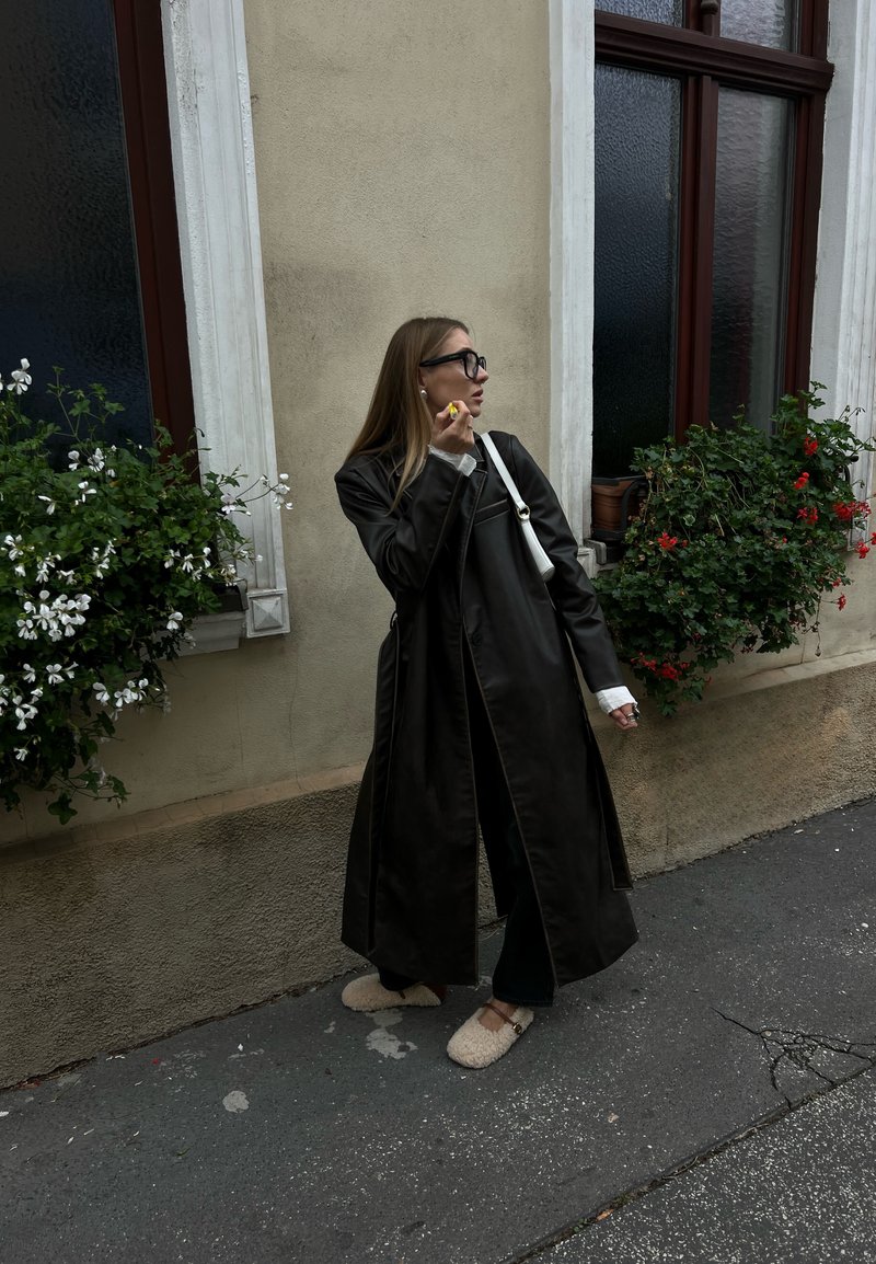 Μαύρο δερμάτινο μακρύ παλτό με δομημένο σχέδιο, οι μανσέτες του λευκού πουκαμίσου ορατές, και μπεζ βελούδινα παντόφλες; φλοράλ πράσινες γλάστρες στο φόντο.