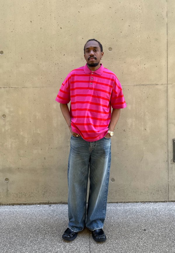 Homme aux cheveux courts et à la barbe, portant un polo rayé rose et rouge, un jean large bleu et des chaussures noires, debout contre un mur en béton.