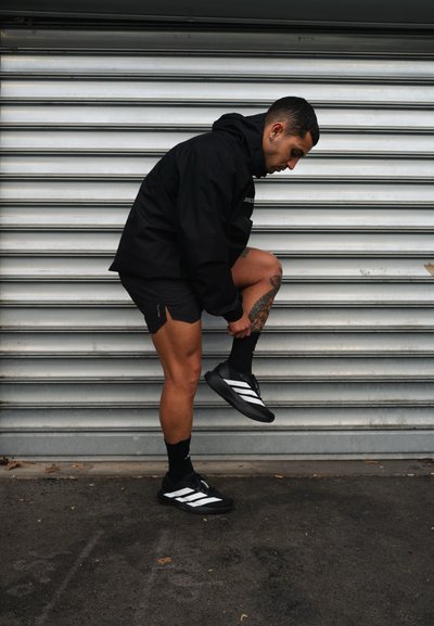 Hombre con ropa deportiva negra ajustándose un calcetín junto a una pared de persianas metálicas, mostrando una pierna tatuada y zapatillas negras con rayas blancas.
