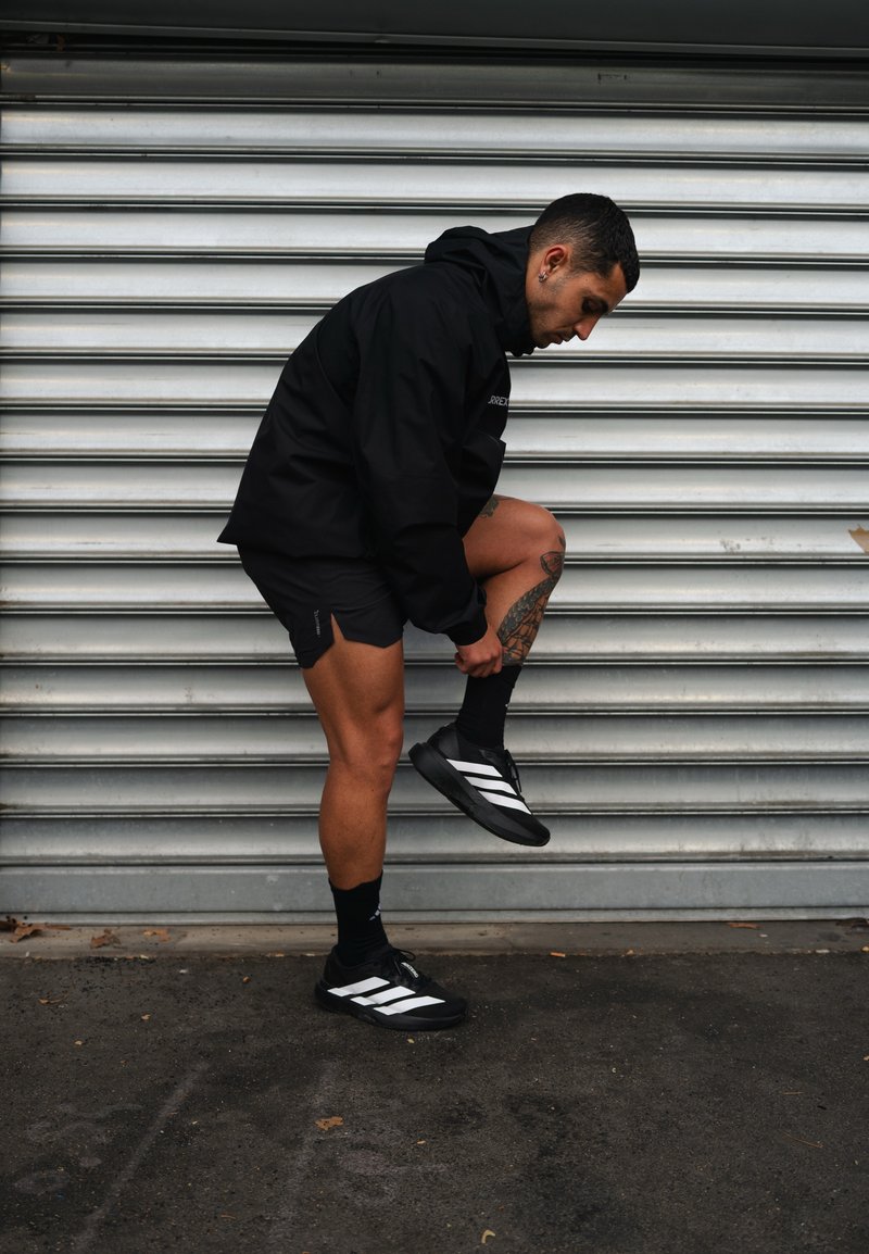 Homme en tenue de sport noire ajustant sa chaussette près d'un mur de shutter en métal, montrant une jambe tatouée et des baskets noires avec des rayures blanches.