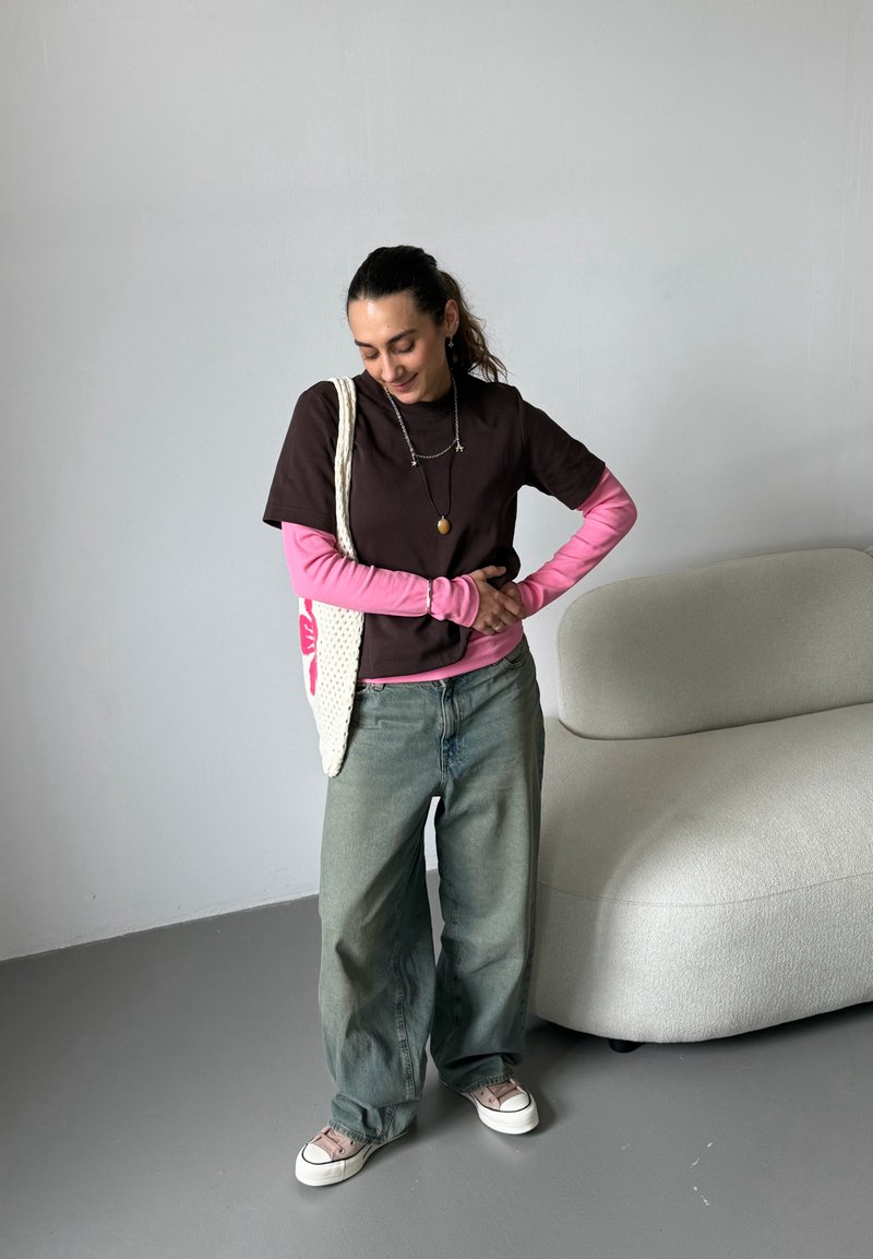 Junge Frau, die geschichtete rosa und braune Oberteile mit lockeren Jeans und Turnschuhen trägt, steht neben einem weißen strukturierten Sofa und trägt eine weiße Einkaufstasche.