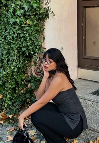 Grey strapless top with textured fabric, high-waisted black trousers, long wavy hair, and round glasses, seated against a green ivy backdrop.