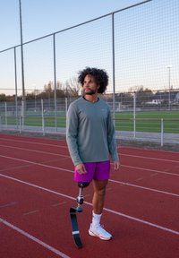 Mannlig idrettsutøver med en løpsprotese står på en rød løpebane iført lilla shorts og en grå genser med lange ermer, ser mot høyre.