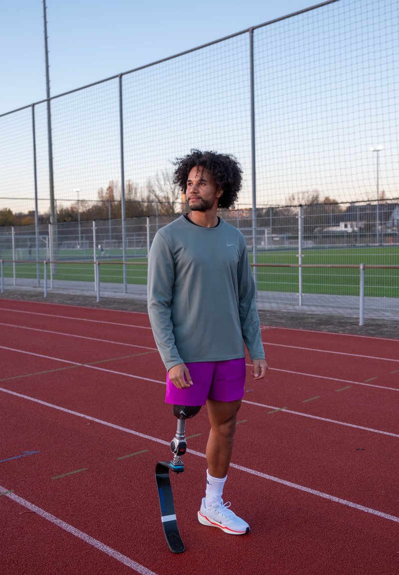 Atleta masculino com prótese de lâmina a correr, está numa pista vermelha, veste calções roxos e uma camisa cinza de manga comprida, olhando para a direita.