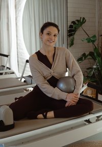 Mladá žena v cvičebnom oblečení sedí v lotosovom sede na Pilates reformeri, drží sivú cvičebnú loptičku, so zelenými rastlinami a závesmi v pozadí.