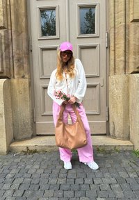Mujer con un top blanco texturizado y pantalones a rayas rosas, sosteniendo un ramo de flores, con un bolso de bronce brillante y llevando una gorra rosa y gafas de sol.