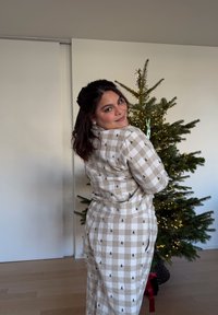 Bézs és fehér kockás ruha apró fa mintákkal, laza szabással és puha tapintással. A háttérben egy feldíszített karácsonyfa látható.