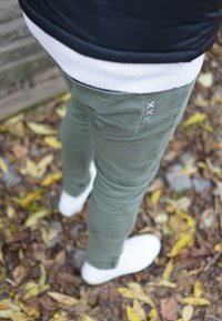 Slim-fit olijfgroene broek met decoratieve stiksels op het achterzak, gecombineerd met witte sneakers, tegen een achtergrond van bladeren.