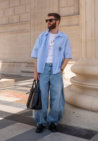 Hombre con barba y gafas de sol viste una camisa azul claro sobre camiseta blanca y jeans holgados, sostiene una bolsa negra cerca de una columna de piedra al aire libre.