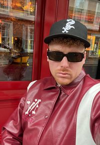 Giacca di pelle bordeaux e bianca con un logo. Cappellino nero con un logo bianco. Occhiali da sole scuri. Lo sfondo mostra una porta rossa e riflessi.