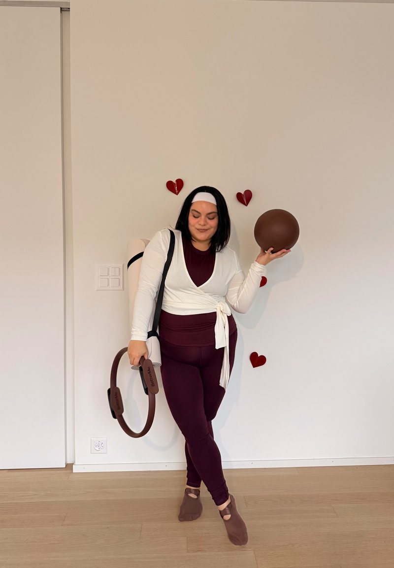 Femme en tenue de sport bordeaux et blanche tenant un anneau de Pilates et une balle, debout pieds nus sur un sol en bois devant un mur uni.