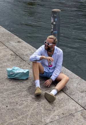 Camisa azul claro, pantalones cortos beige y calzado marrón. Sentado en el concreto junto al agua con una bolsa color aqua y una bebida rosa en la mano. Las gafas de sol complementan el look.