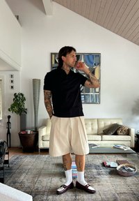 Man met tatoeages, gekleed in een zwart shirt, beige shorts, witte sokken en bruine schoenen, staande in een woonkamer met een crème bank en abstracte wandkunst.