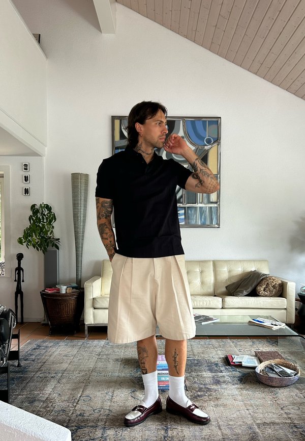 Man with tattoos wearing black shirt, beige shorts, white socks, and brown shoes standing in a living room with a cream sofa and abstract wall art.