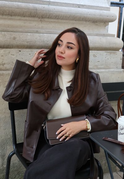 Mujer con largo cabello castaño, vestida con una chaqueta de cuero marrón y una blusa blanca, sentada en una mesa de café, sosteniendo un bolso marrón a juego y tocándose el cabello.
