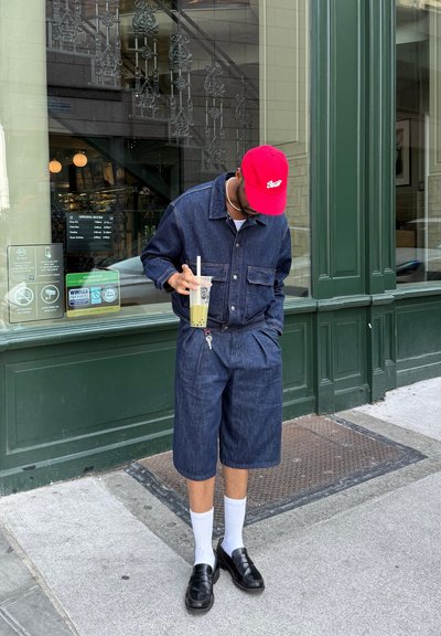 Chaqueta de mezclilla y pantalones cortos en azul oscuro, tejido texturizado. Gorra roja, calcetines blancos y zapatos negros. Sostenido una taza con una pajita. Fondo verde.