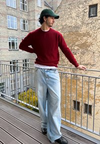 Mees, kes kannab burgundi tooni kootud kampsunit, valget särki ning helesiniseid laiade säärtega pükse, seisab rõdul, peas roheline nokamüts ja jalas mustad jalanõud.