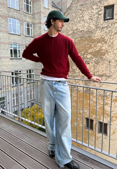 Hombre con suéter de punto burdeos, camisa blanca y vaqueros de pierna ancha azul claro, de pie en un balcón con gorra verde y zapatos negros.