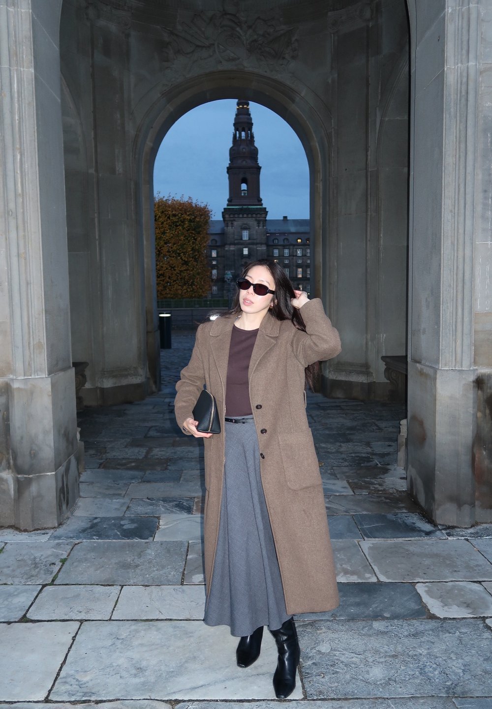 Brauner Wollmantel, grauer Plissée-Rock, schwarze Leder-Stiefel bis zum Knie und dunkle Sonnenbrille. Bogen mit Steintextur im Hintergrund.
