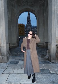 Brauner Wollmantel, grauer Plissée-Rock, schwarze Leder-Stiefel bis zum Knie und dunkle Sonnenbrille. Bogen mit Steintextur im Hintergrund.