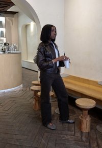 Braune Lederjacke, weißes Oberteil, schwarze Hose, schwarz-weiße gemusterte Ballerinas, stehend mit einem Getränk in der Nähe von hellen Holzmöbeln in einem Café.