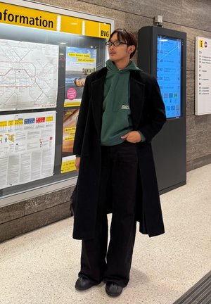 Persona con un abrigo largo negro sobre una sudadera verde con capucha, pantalones anchos negros y zapatos negros. De pie cerca de un panel de información de transporte público.