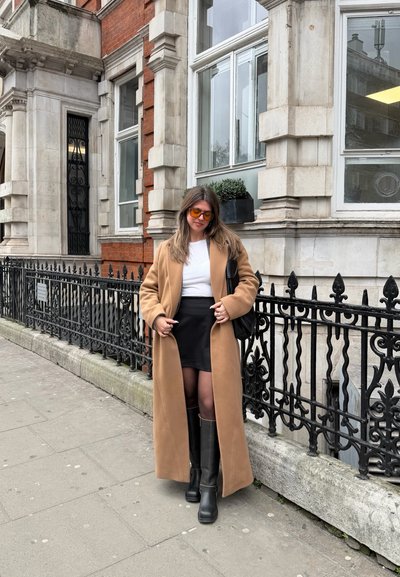Mujer con un abrigo largo camel, falda negra, botas negras y gafas de sol naranjas de pie en la acera de una ciudad junto a una reja de hierro ornamentada.