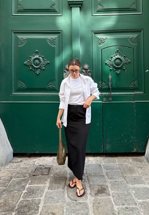Camisa blanca de botones sobre una camiseta blanca, falda lápiz negra, chanclas, sosteniendo un bolso marrón, frente a una puerta verde ornamentada.