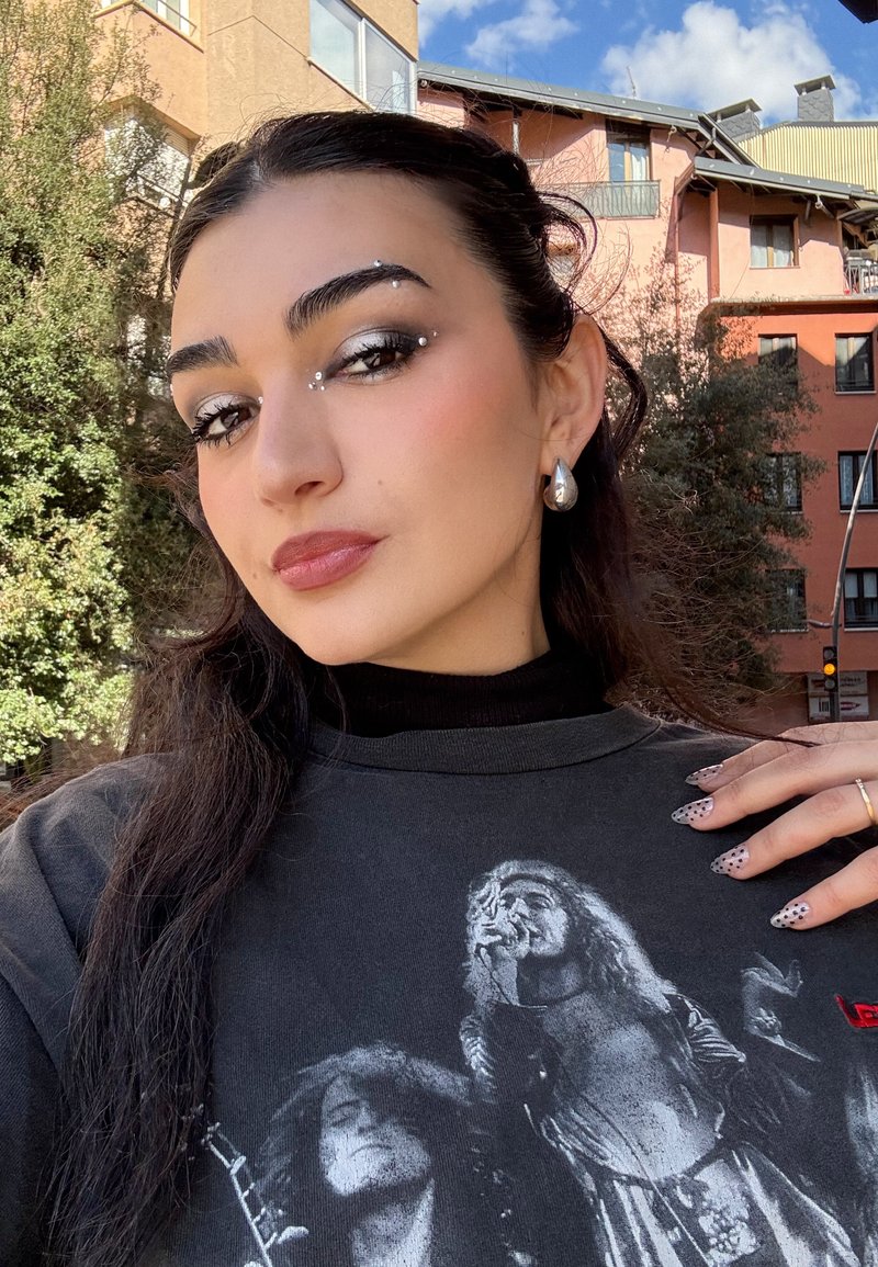 Jeune femme à l'extérieur avec des bijoux d'argent pour les yeux, des boucles d'oreilles en forme de goutte et un t-shirt noir présentant une photo vintage d'un groupe de rock.