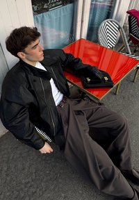 Veste en cuir noire avec une chemise blanche, un pantalon foncé et des gants noirs. Sac noir avec ferrures dorées posé sur une table rouge à côté d'une chaise chevron.