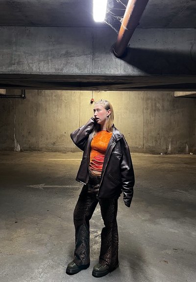 Mujer de pie en un aparcamiento subterráneo de hormigón con poca luz, vistiendo una chaqueta de cuero negra, un top naranja texturizado y pantalones oscuros con estampado.