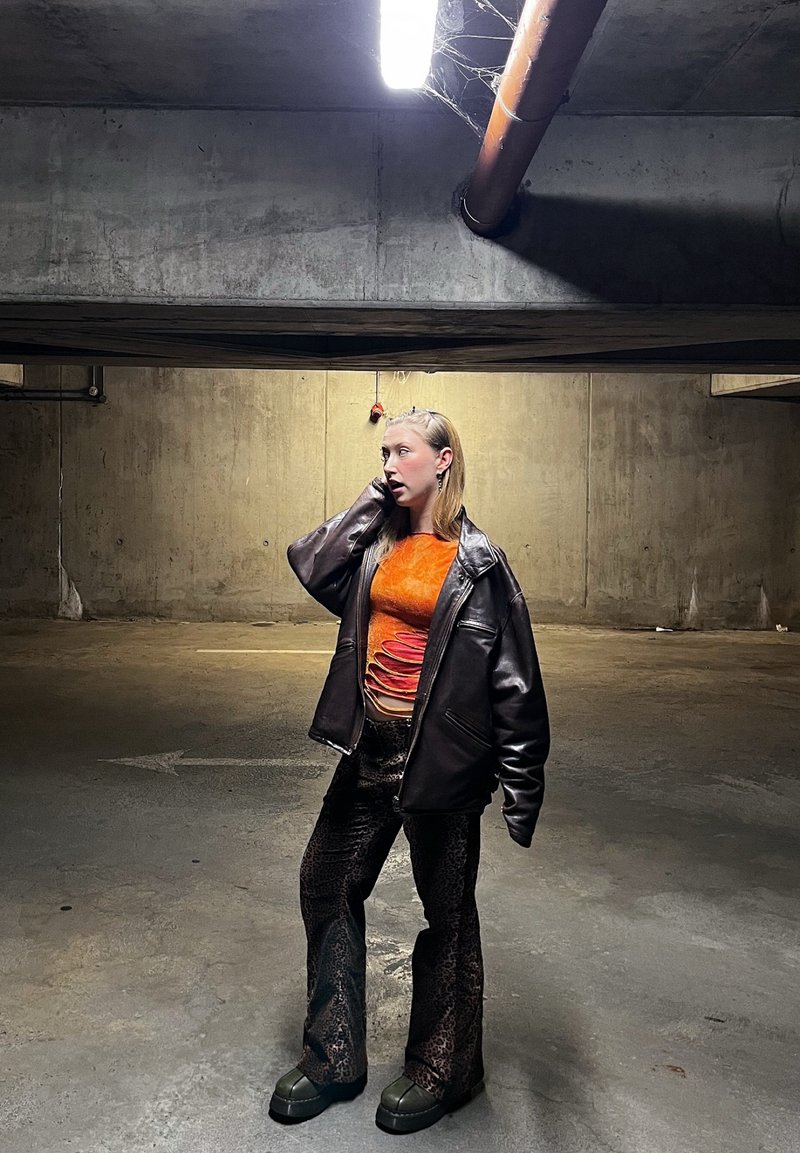 Femme debout dans un parking en béton faiblement éclairé, portant une veste en cuir noire, un haut orange texturé et un pantalon sombre à motifs.