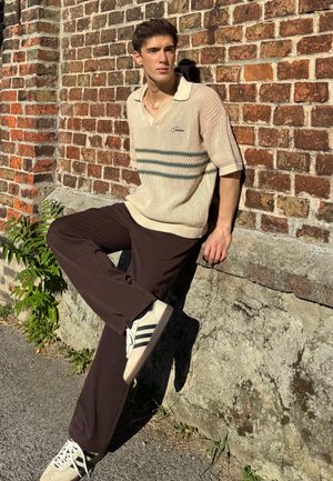Beige gebreide polo met groene strepen, bruine broek en witte sneakers met zwarte accenten. Staand tegen een bakstenen muur.