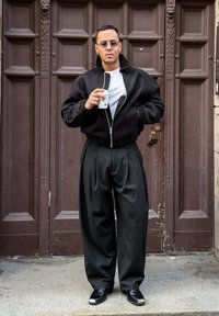 Crna bomber jakna s visokim ovratnikom, bijela majica, crne hlače s naborima i crne cipele sa srebrnim detaljem. Drži papirnatu šalicu.