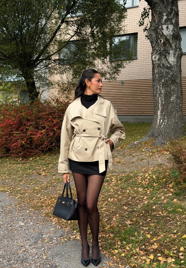 Mujer con abrigo beige con cinturón, vestido negro y medias caminando al aire libre por un camino con hojas, sosteniendo un bolso negro cerca de árboles y un edificio de ladrillo.