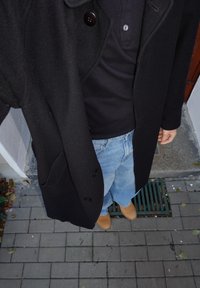 Musta villatakki napilliseen kaulukseen, päällä tumma poolopaita, yhdistetty vaaleansiniin väljähköihin farkkuihin ja vaaleanruskeisiin puukenkään asfalttipinnalla.