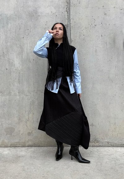 Mujer con el pelo largo trenzado, vestida con una camisa azul, chaleco negro, falda negra asimétrica y botas negras de tacón, apoyada contra una pared de hormigón.