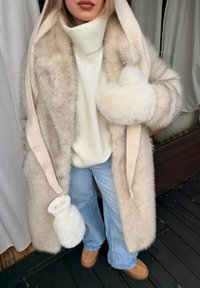 Femme portant un manteau en fourrure beige, un pull à col roulé crème, un jean bleu clair et des chaussures tans, debout sur un sol en bois foncé.