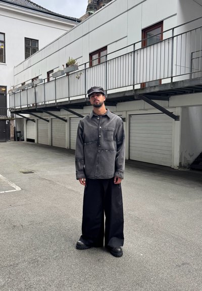 Camisa gris de botones con bolsillo frontal y costuras en contraste, combinada con pantalones anchos negros y una boina, con un fondo urbano.