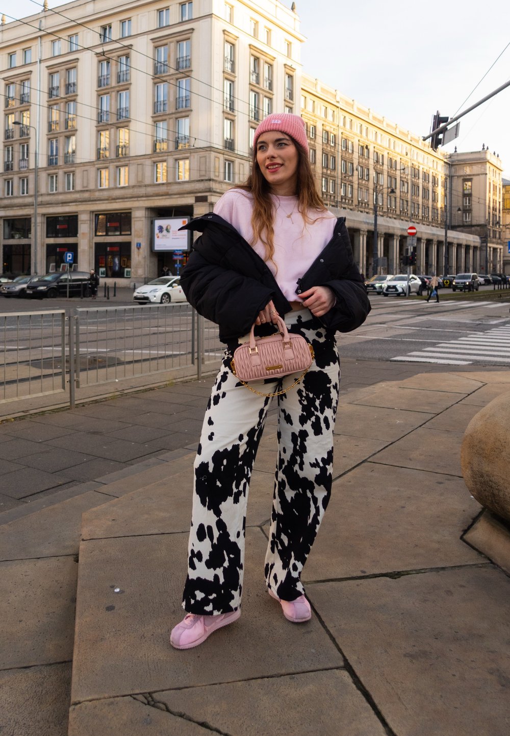 Pantalon large en imprimé vache noir et blanc, sweat-shirt rose, doudoune noire et sac à main rose pâle. Debout sur un trottoir de la ville.