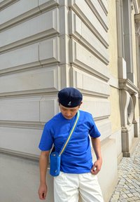 T-shirt di cotone blu, maniche corte. Borsa a tracolla gialla e blu, tessuto testurizzato. Pantaloni chiari; in piedi vicino a un muro testurizzato.